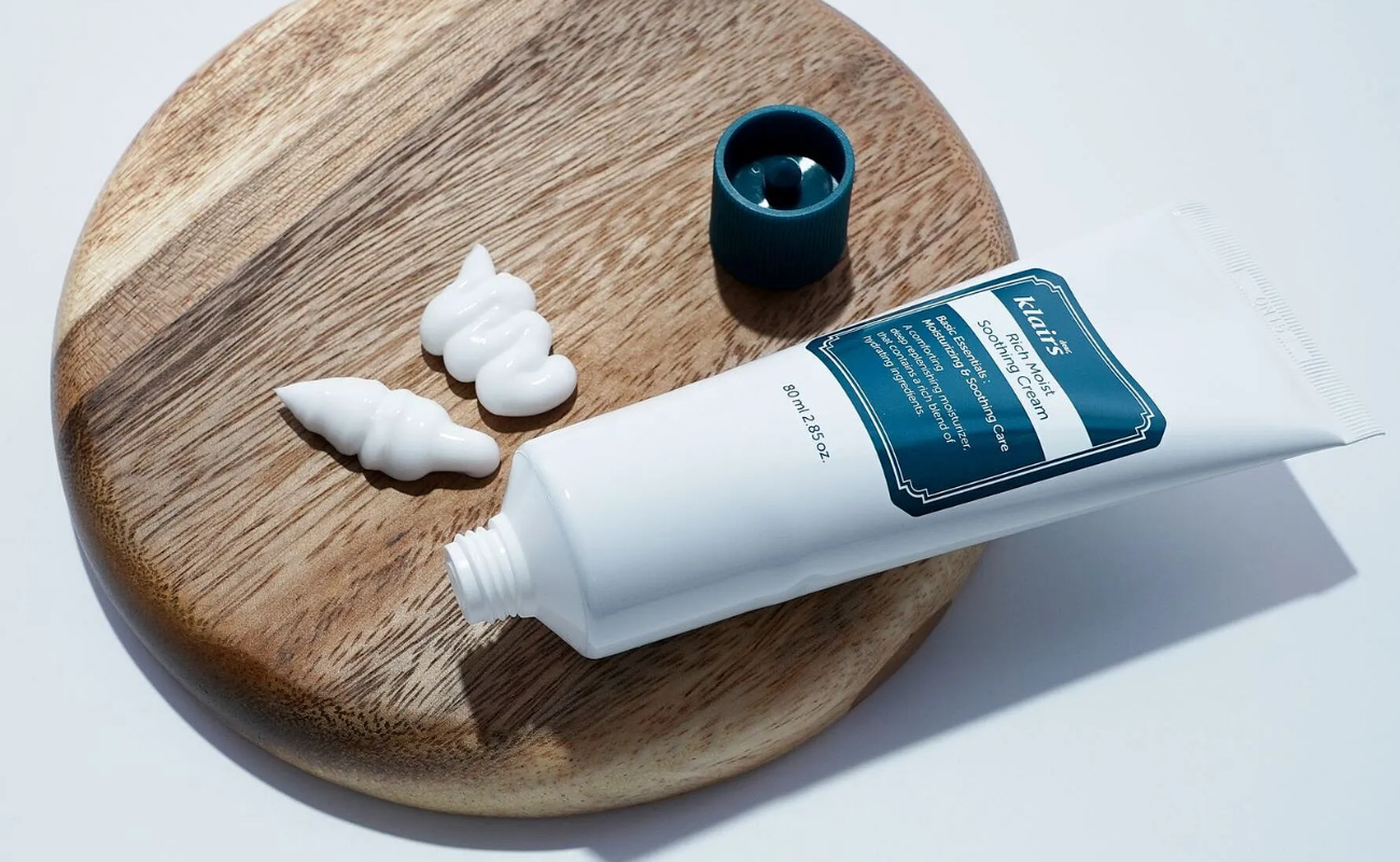 Närbild på Klairs Rich Moist Soothing Cream-tub och klickar av vit ansiktskräm på ett träfat.